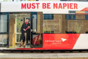 Graduate standing on an Edinburgh tram branded with ‘Must Be Napier’ and the Edinburgh Napier University logo, celebrating graduation day