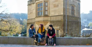 Three students sit in front of a historic building on Bangor University campus.